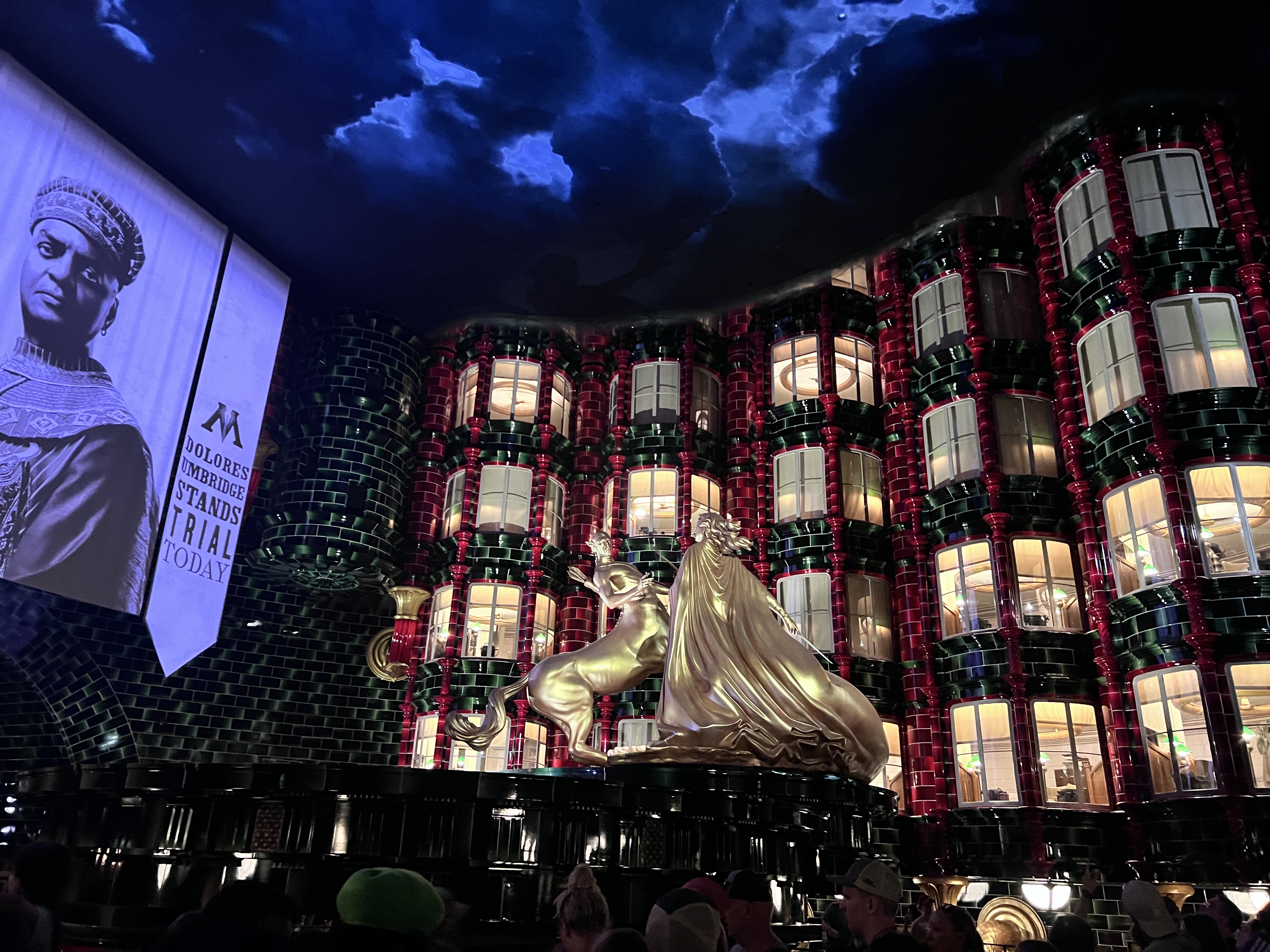 The Ministry of Magic atrium queue: the golden Fountain of Magical Brethren rises beneath a stormy enchanted ceiling, flanked by towering green-and-red tiled columns with glowing office windows, while a banner announces Dolores Umbridge stands trial today